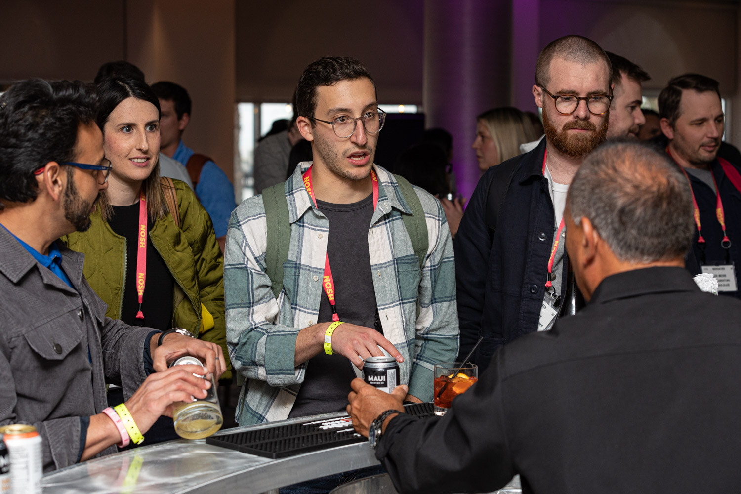 Crowd of attendees with lanyards at a metal bar, a man in a plaid shirt holding a can.
