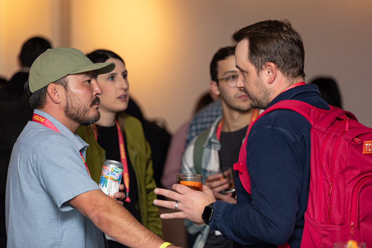 Man in a green cap and light blue shirt talking to a man in a navy sweater with a red backpack, others behind them.