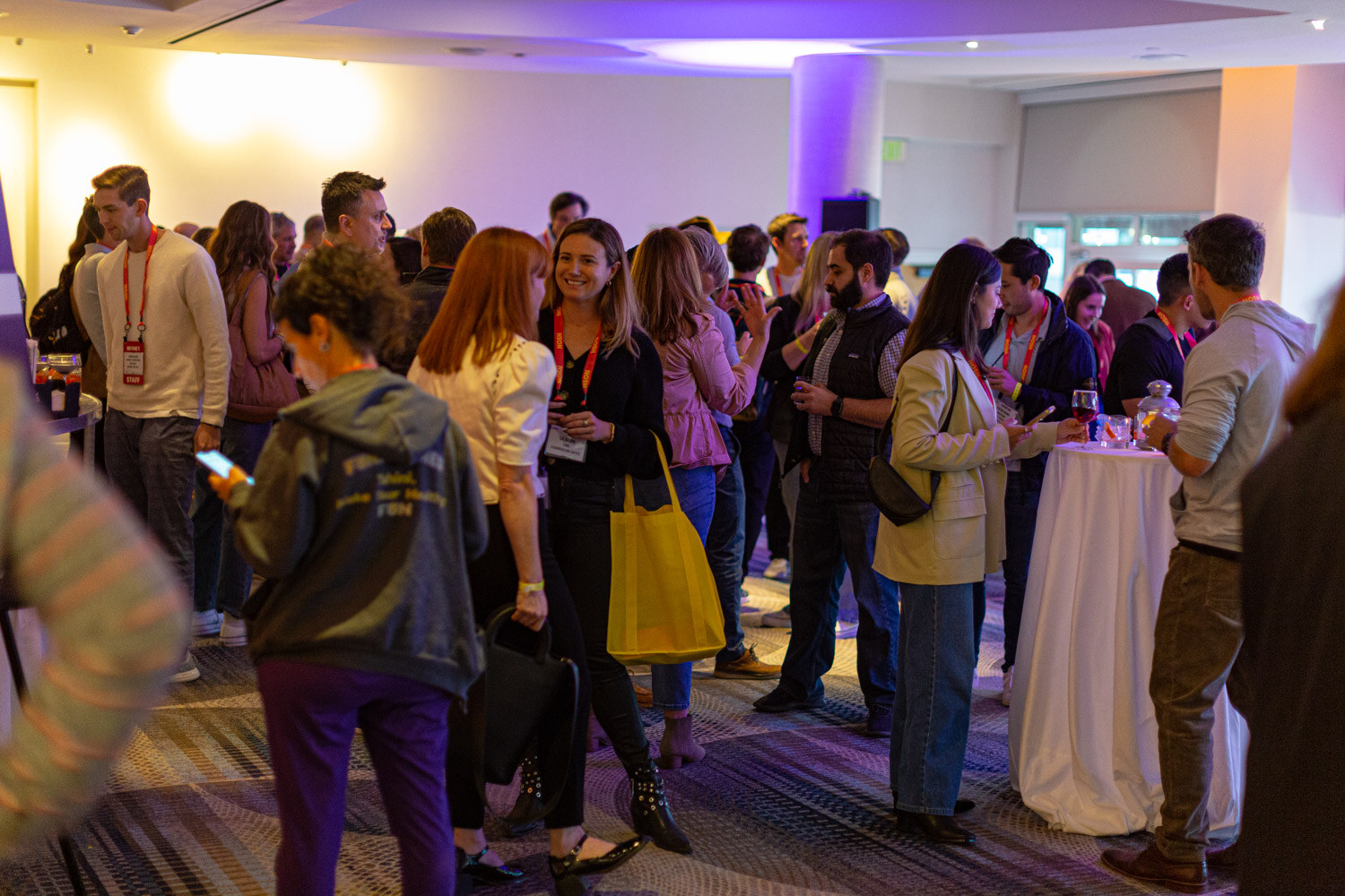 Wide shot of a crowded room of attendees in lanyards standing around cocktail tables.