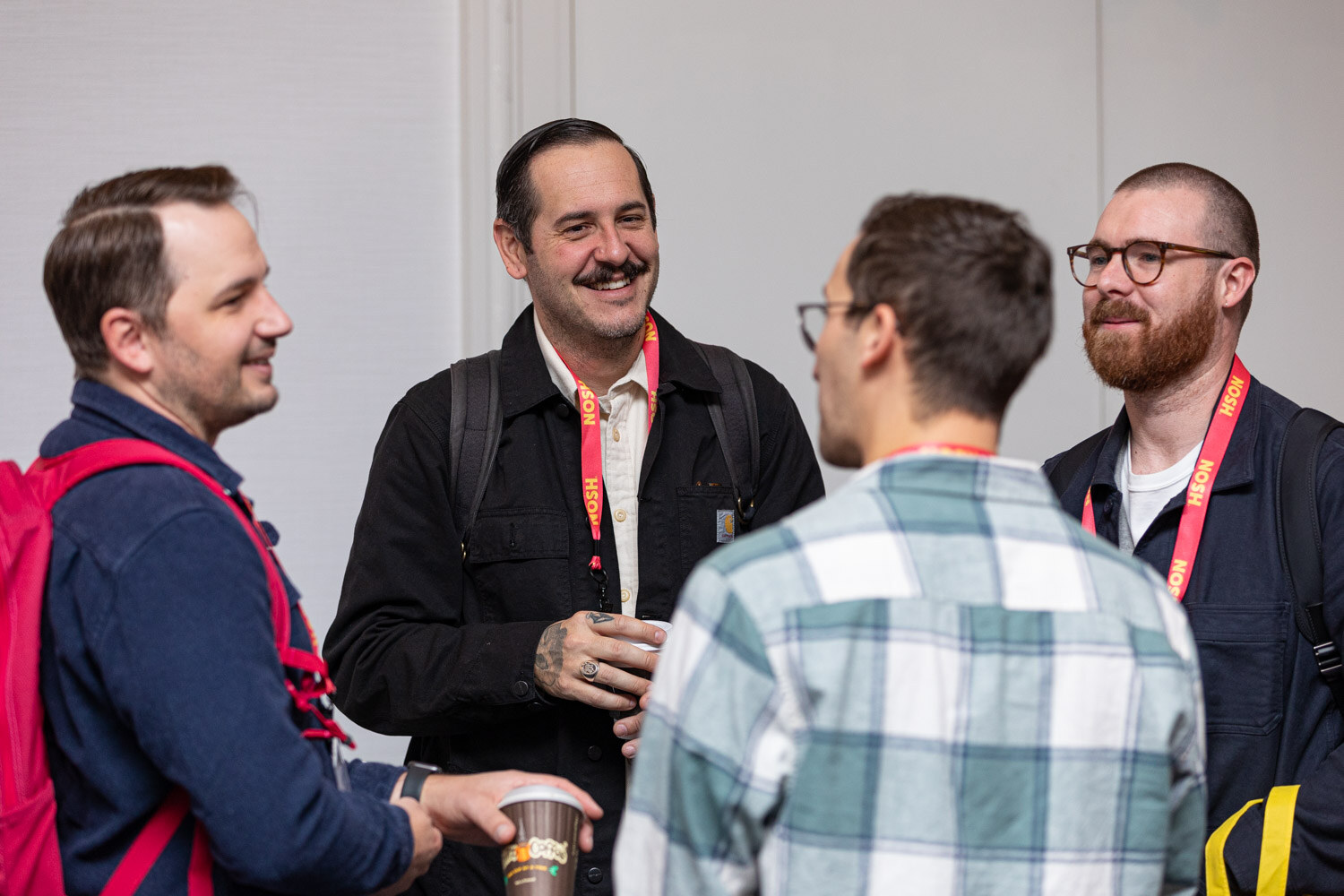 Four men with lanyards standing in a circle, one holding a coffee cup.