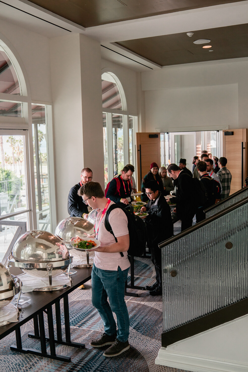 Man with a backpack standing at a buffet table with covered serving dishes, attendees seated beyond a glass railing.