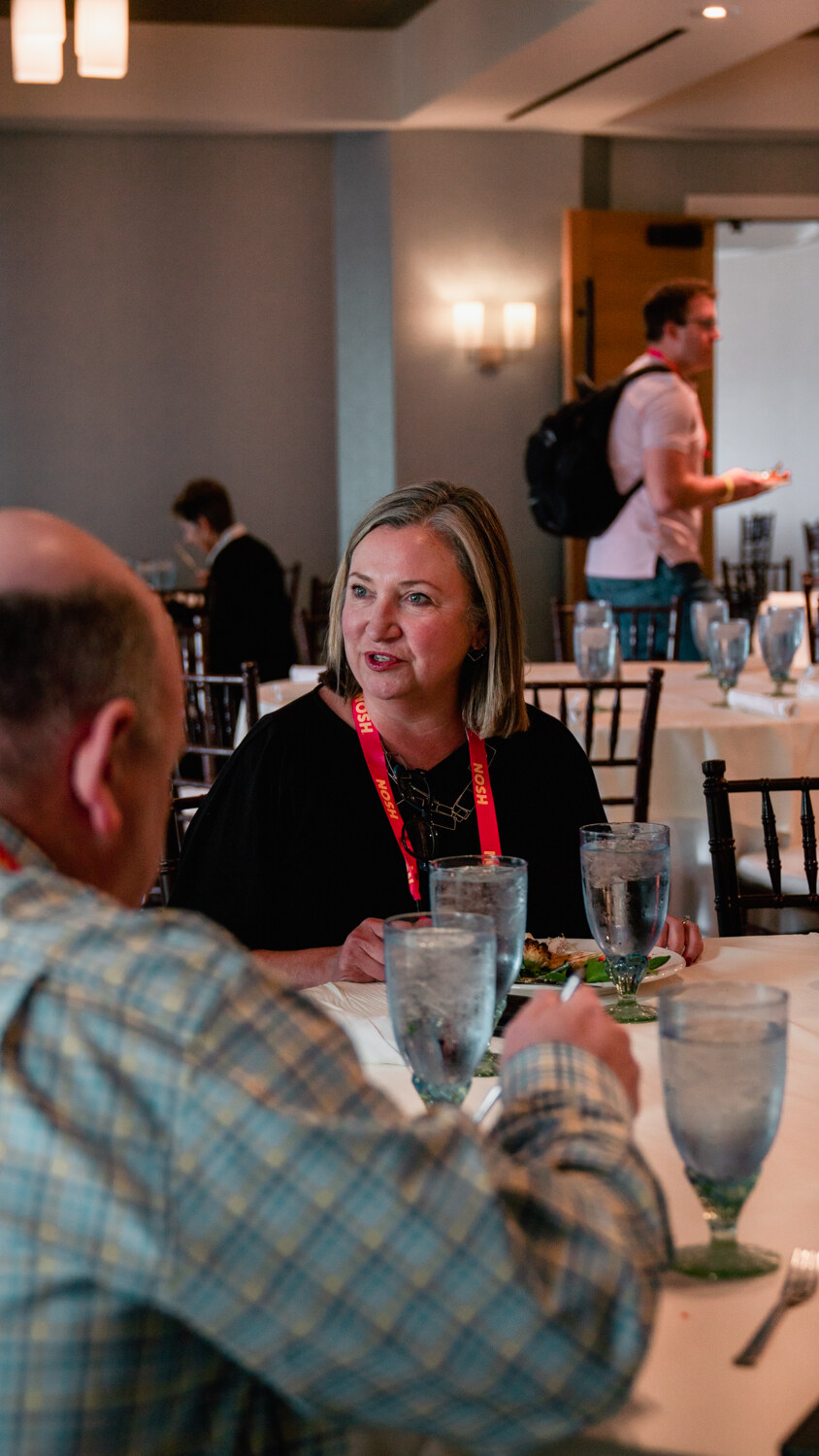 Woman in black with a red lanyard seated at a set table, another man in a plaid shirt with his back to the camera.