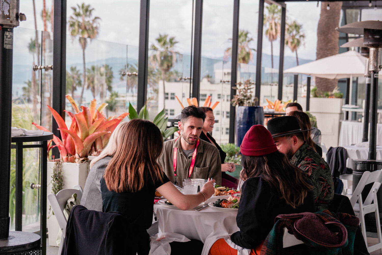 People seated at a table on a glass-walled terrace with palm trees and tall buildings in the background.