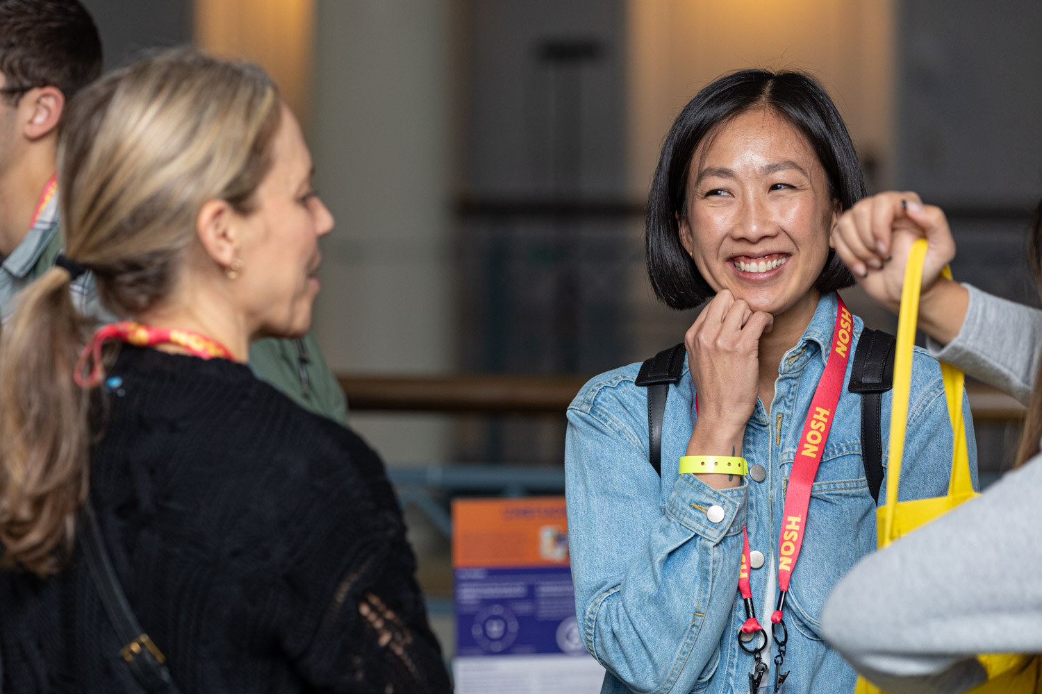 Smiling woman in a denim jacket with a Nosh lanyard resting her chin on her hand.