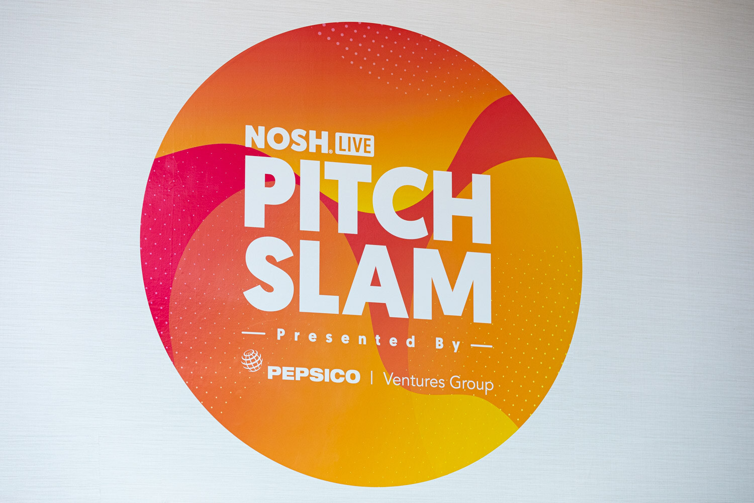 Circular orange and pink sign reading Nosh Live Pitch Slam, presented by Pepsico Ventures Group.