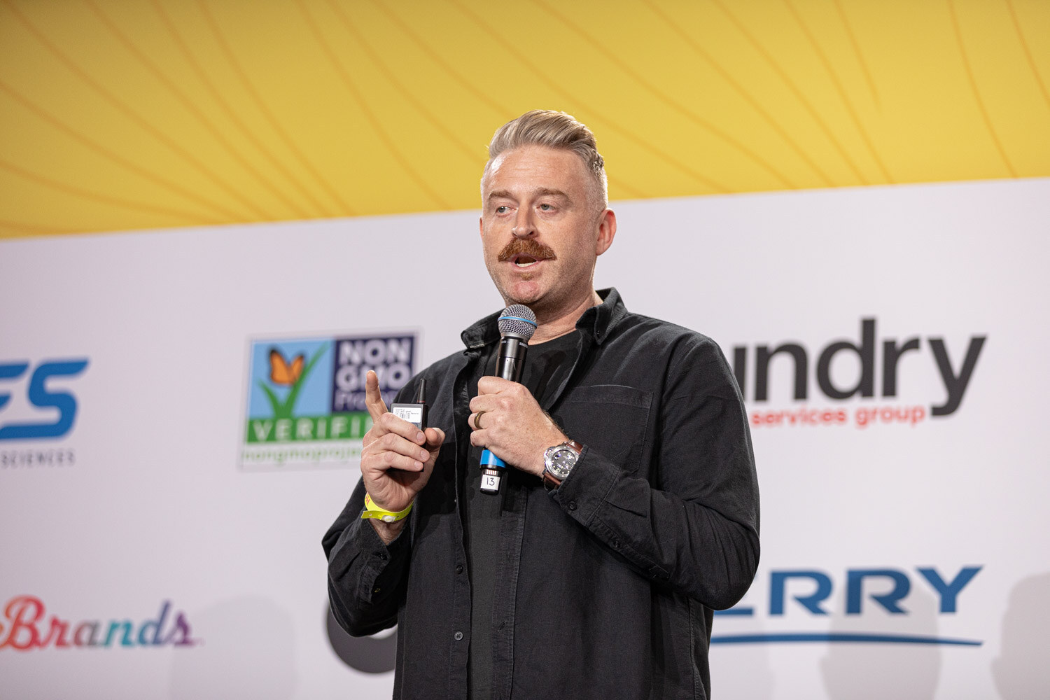 Man with swept-back hair and a mustache holding a microphone in front of a sponsor wall.