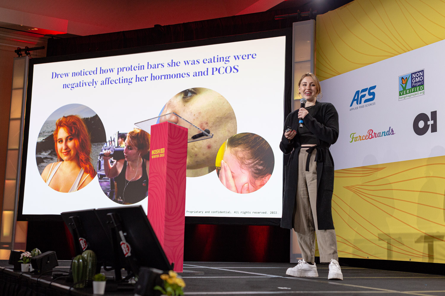 Woman in a dark cardigan standing at the side of a stage with a large slide showing photos and text about protein bars.