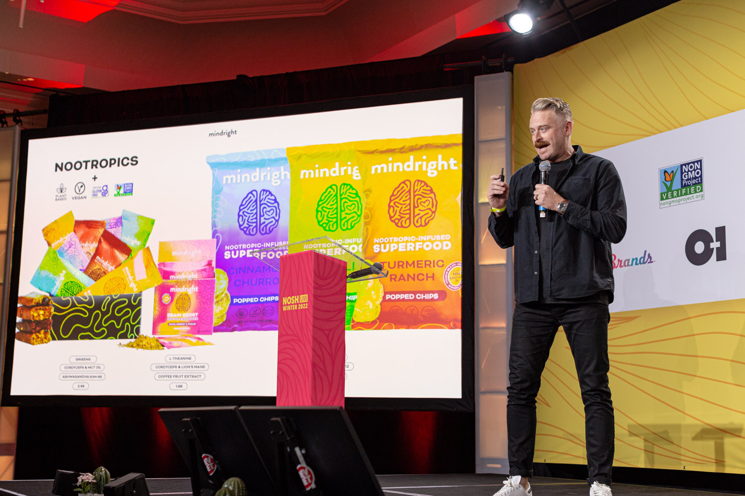 Man in a dark shirt on stage with a large screen showing mindright snack bags.