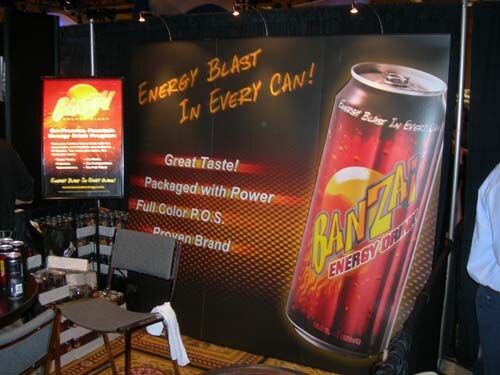 A booth backdrop shows a large Banzai Energy Drink can with the text Energy Blast in Every Can, Great Taste, Packaged with Power, with chairs and a small sign in front.