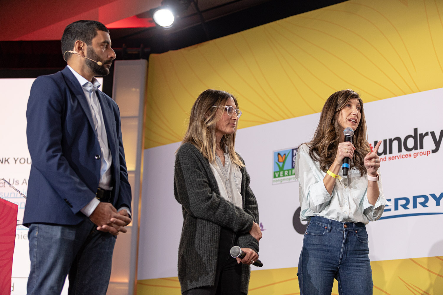 A man in a navy blazer and two women stand on stage, one speaking into a microphone.