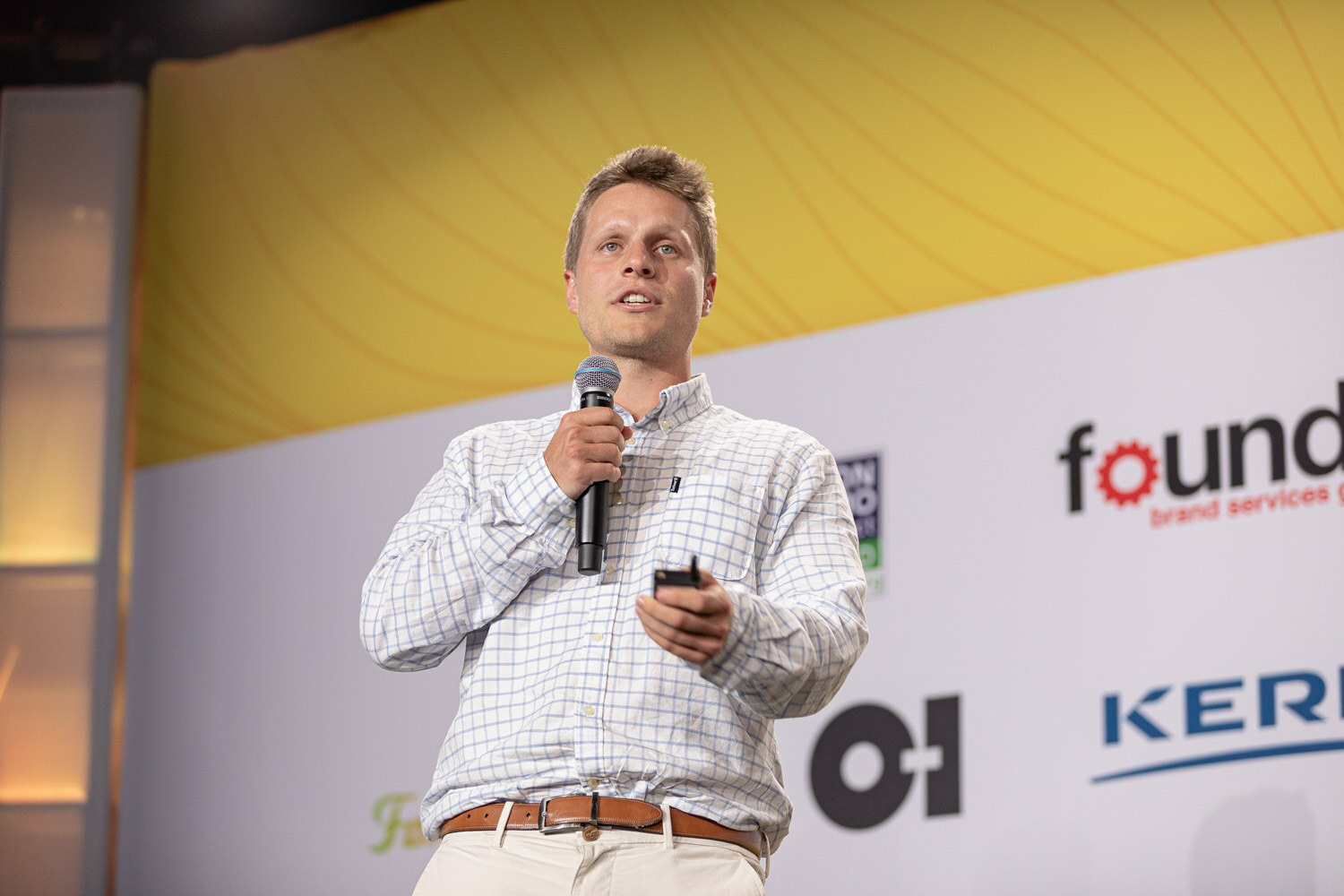 Man in a white checked shirt speaking into a microphone in front of sponsor logos.