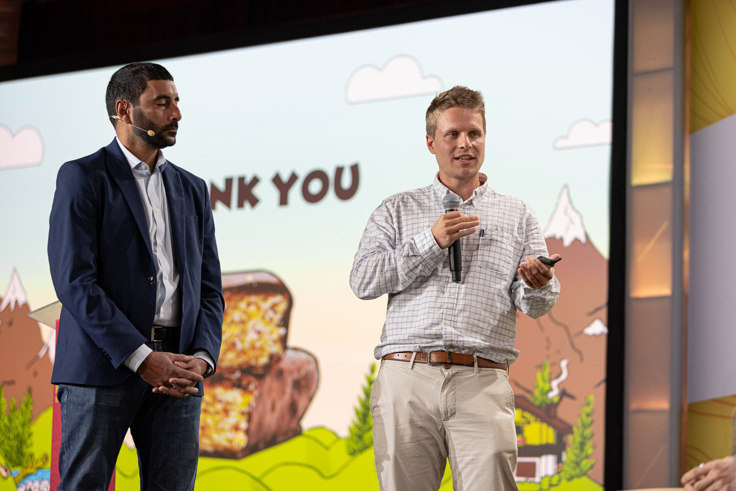 Two men on stage in front of a screen reading Thank You with a cartoon snack bar and mountains.