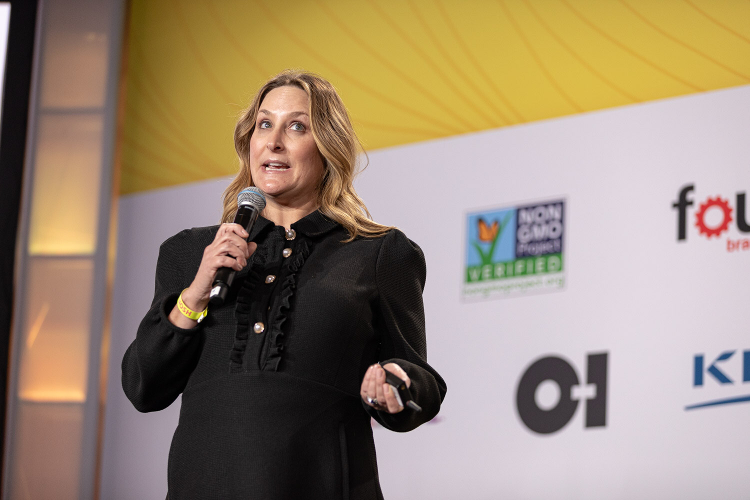 Woman in a black ruffled top holding a microphone in front of a sponsor backdrop.