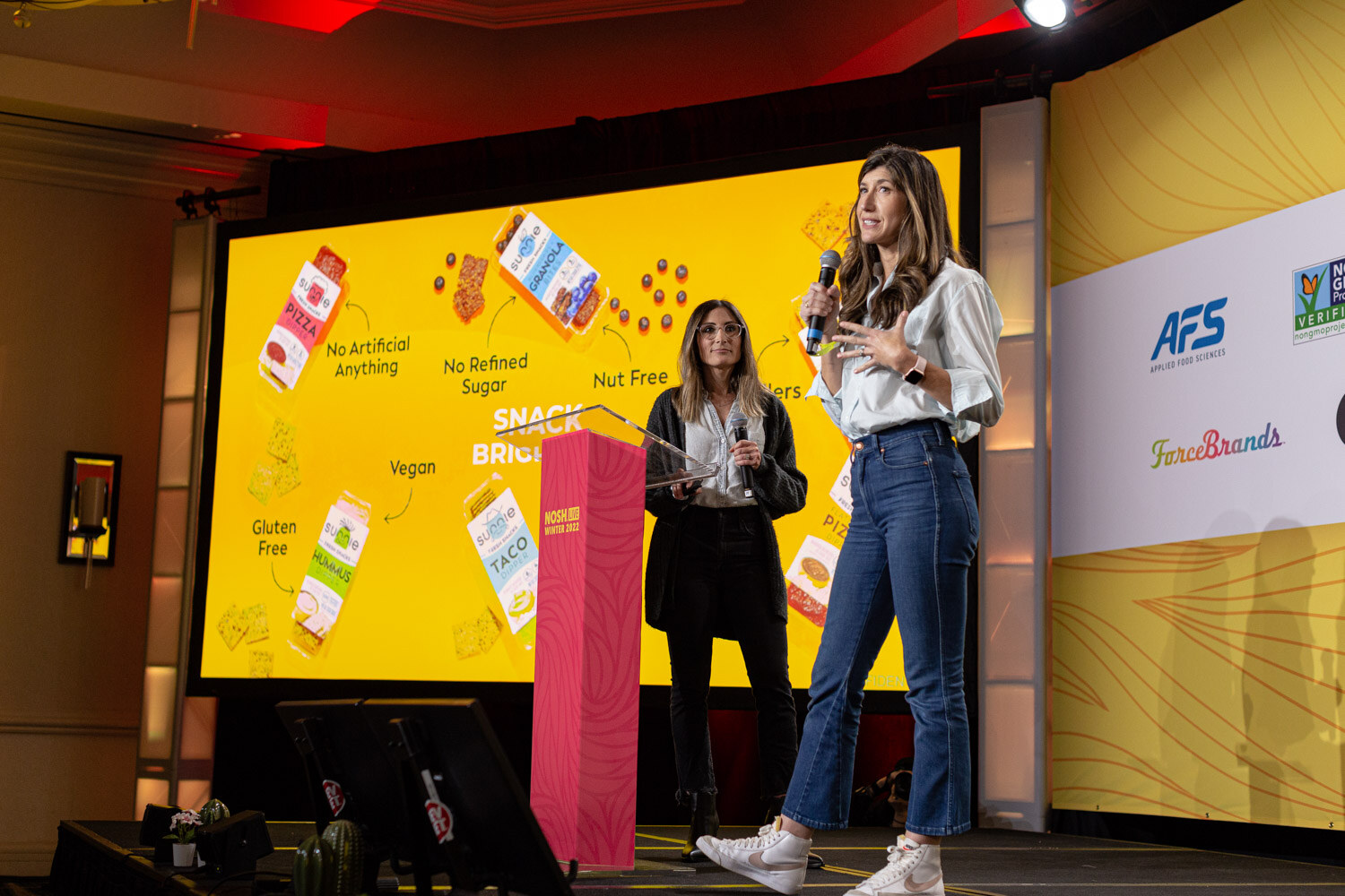 Two women on stage in front of a large yellow screen showing snack packages.