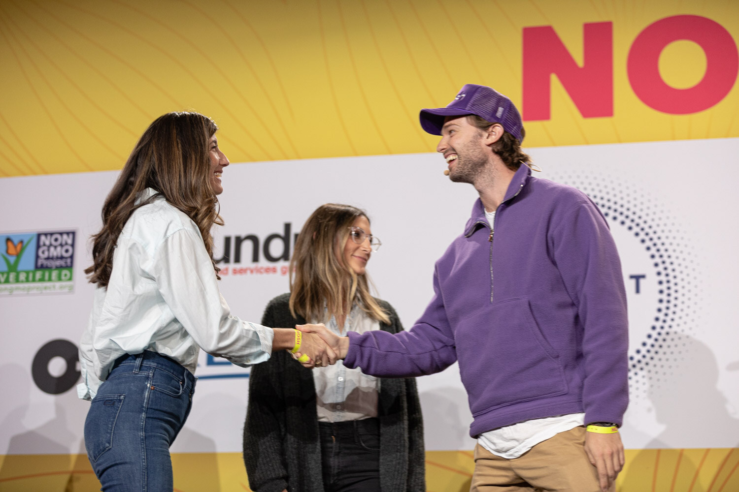 Woman in a light shirt shaking hands with a man in a purple cap and pullover, a woman looking on between them.