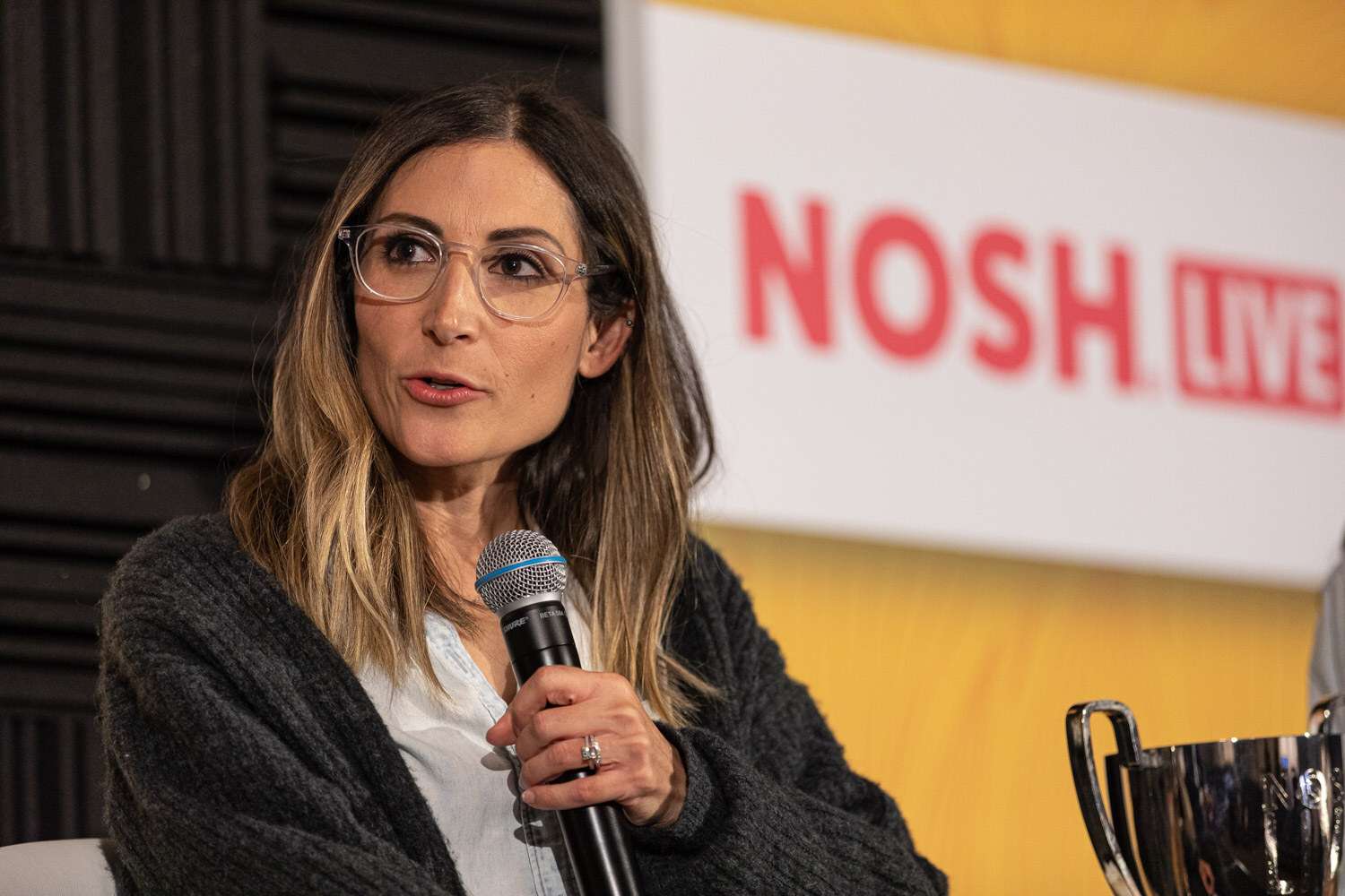 Woman with glasses holding a microphone in front of a Nosh Live sign, a silver trophy at the edge of the frame.