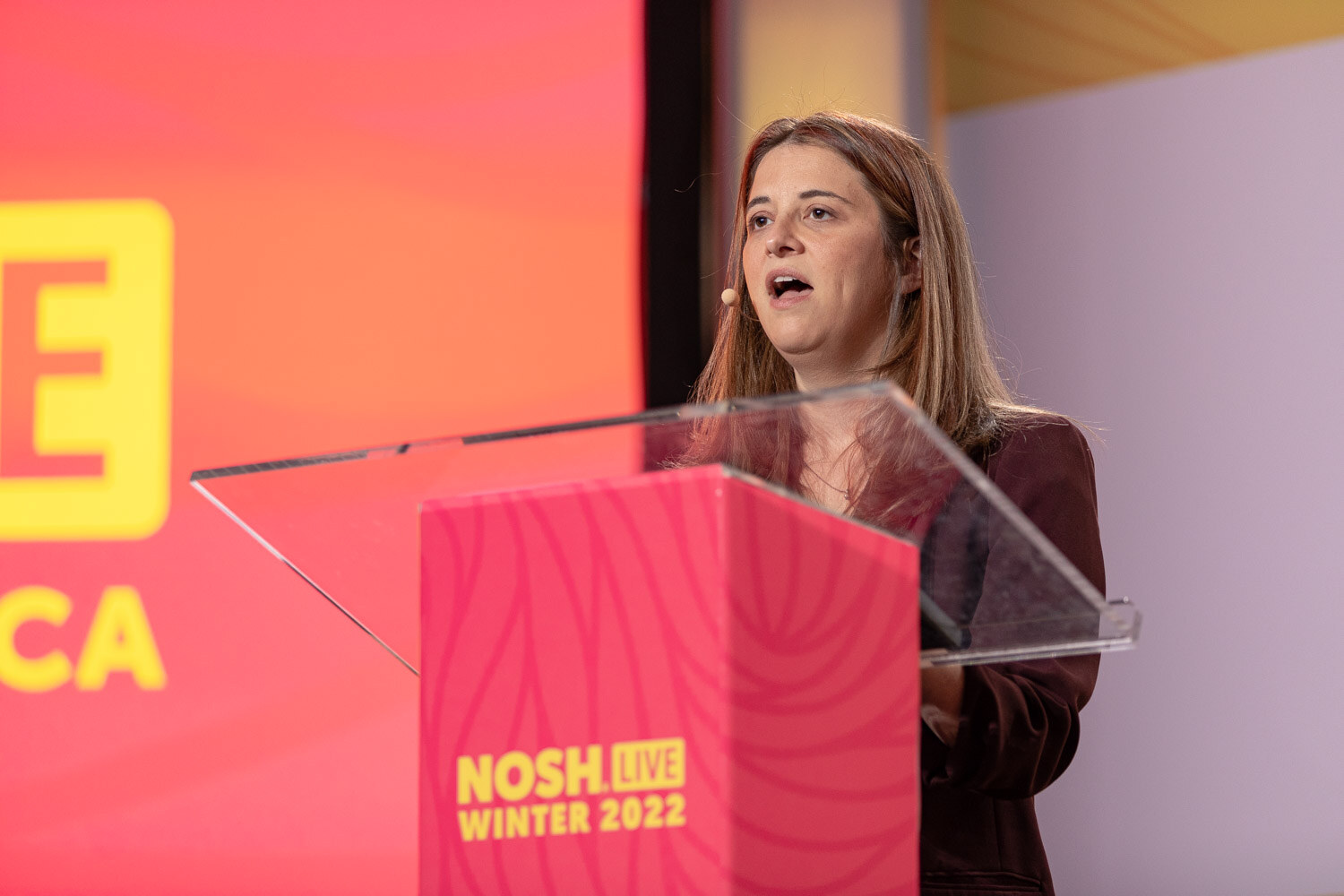 Woman in a burgundy blazer speaking at a pink lectern with a Nosh Live Winter 2022 backdrop.