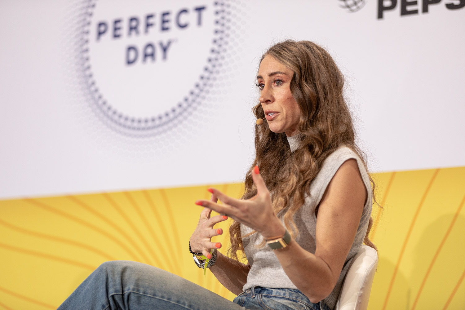 Woman with long wavy hair in a gray sleeveless top gesturing with both hands.