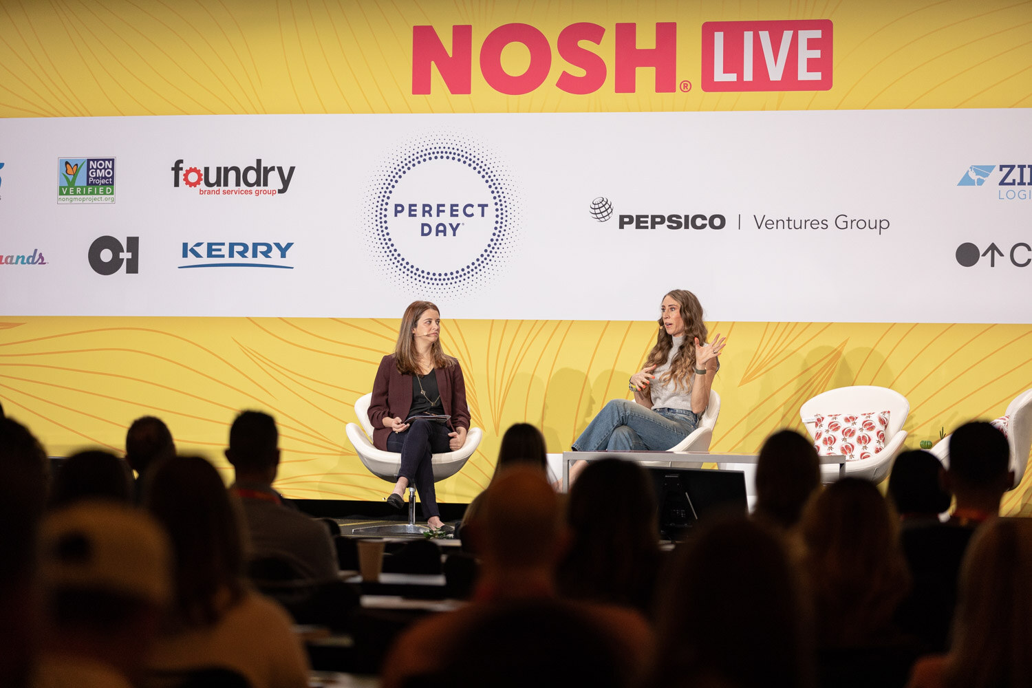 Wide shot with audience silhouettes in the foreground and two seated speakers on a stage with a Nosh Live banner.