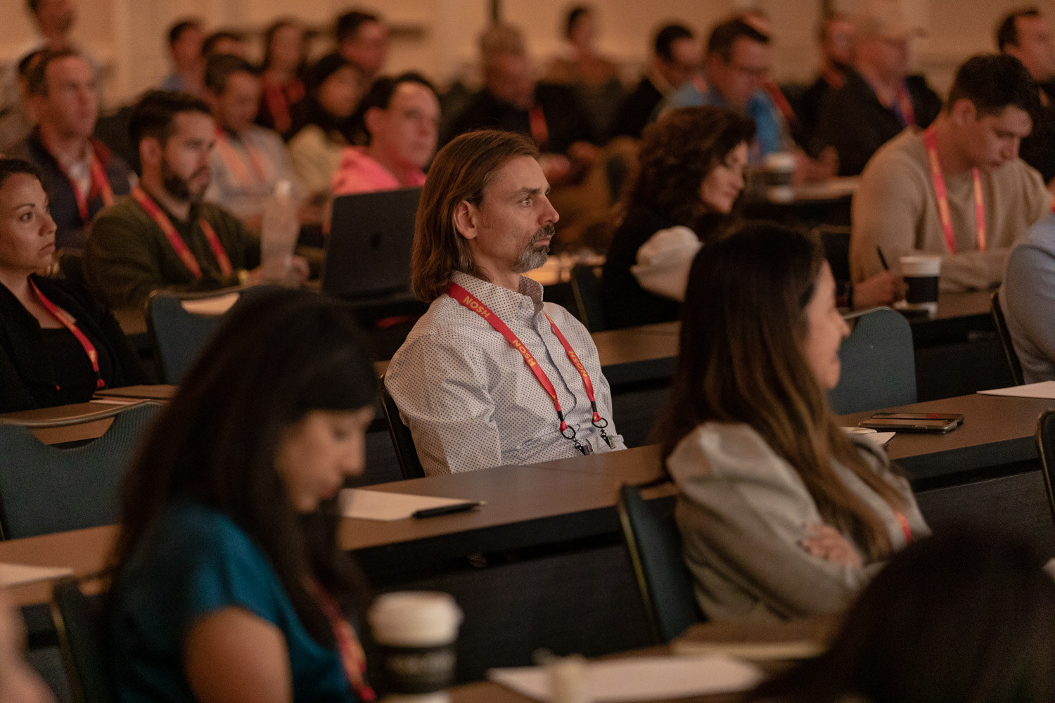 Man with long hair and a lanyard seated among other attendees at classroom-style tables.