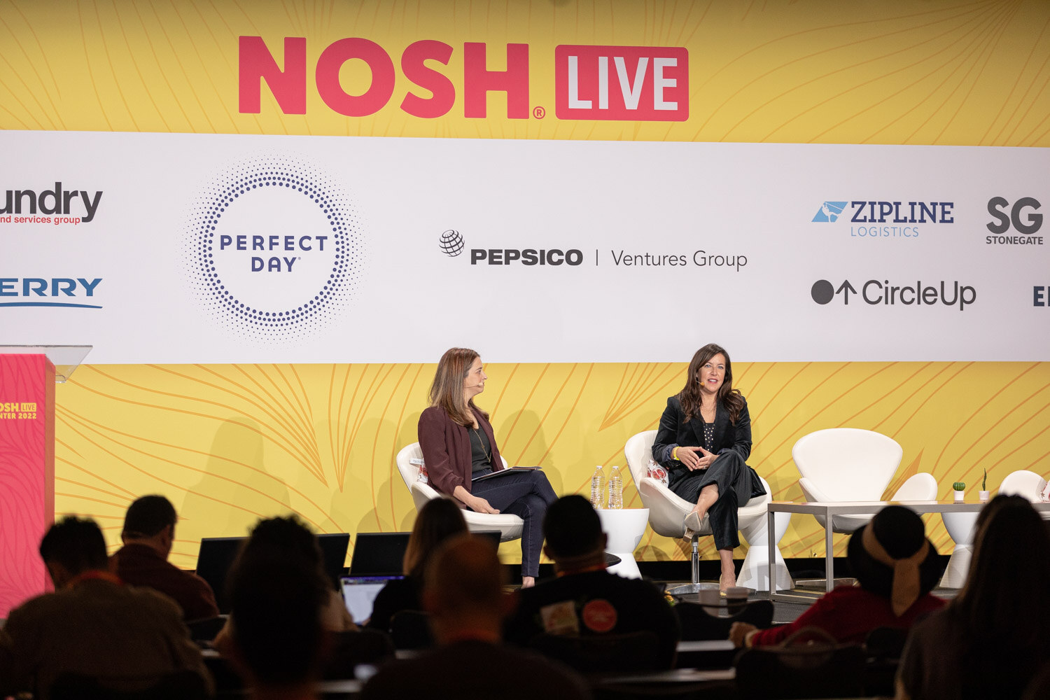Wide shot of two women seated on stage under a Nosh Live banner with audience silhouettes in front.