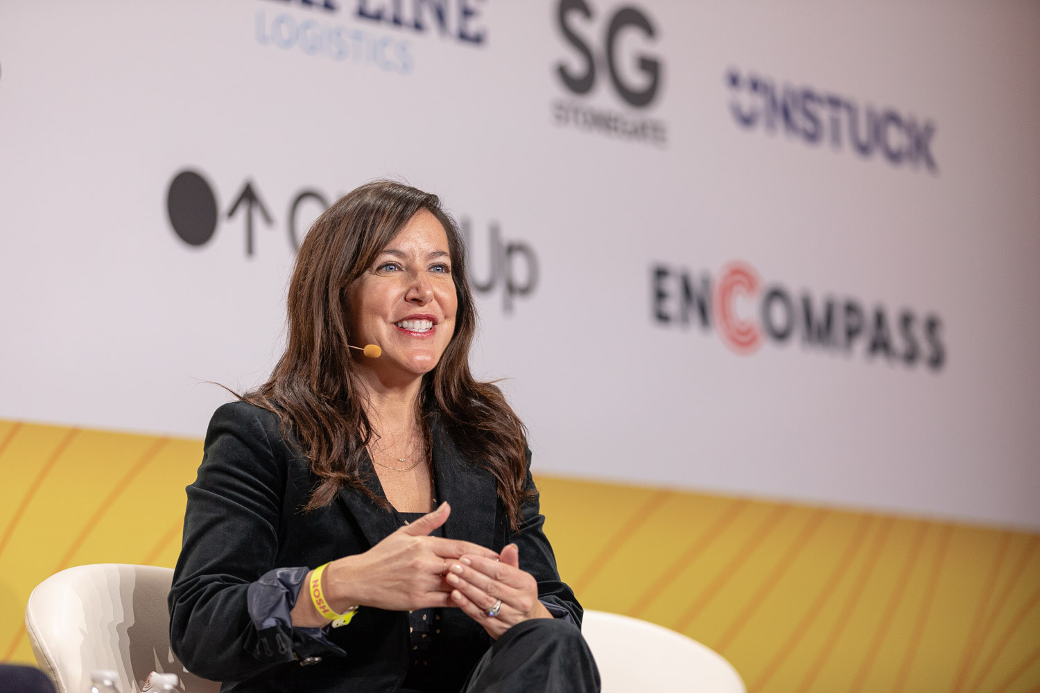 Smiling woman in a dark blazer seated on stage in front of a sponsor backdrop.