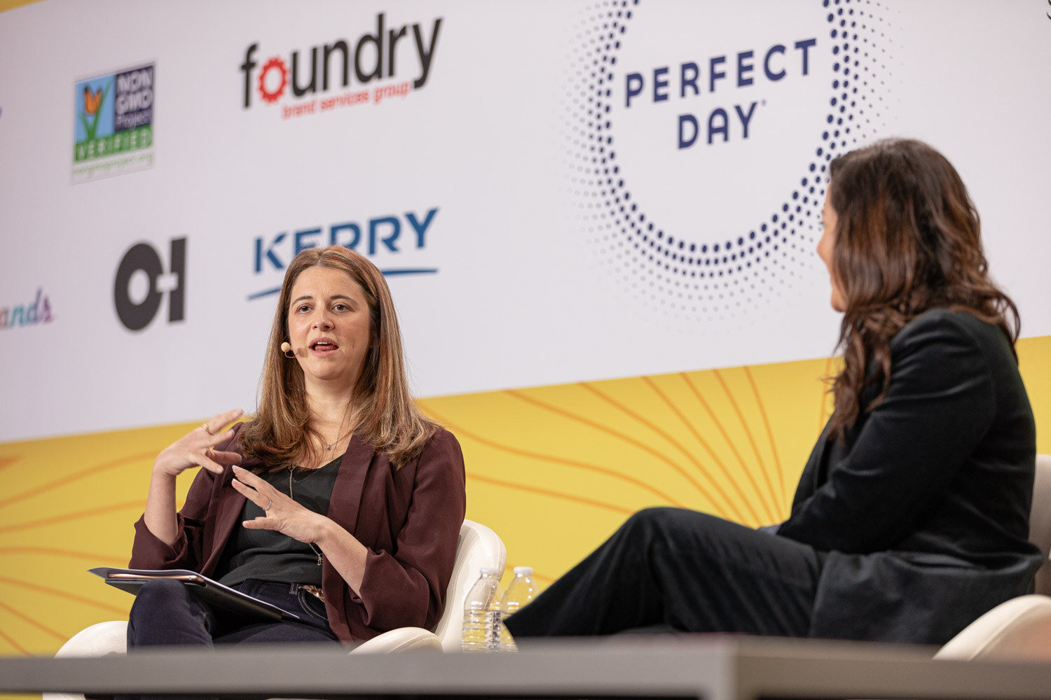 Woman in a burgundy blazer speaking on stage with a second woman seated beside her.