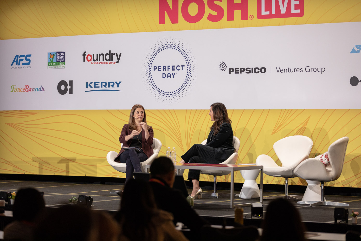 Wide shot of two women on a stage with sponsor logos behind them and the audience in silhouette.