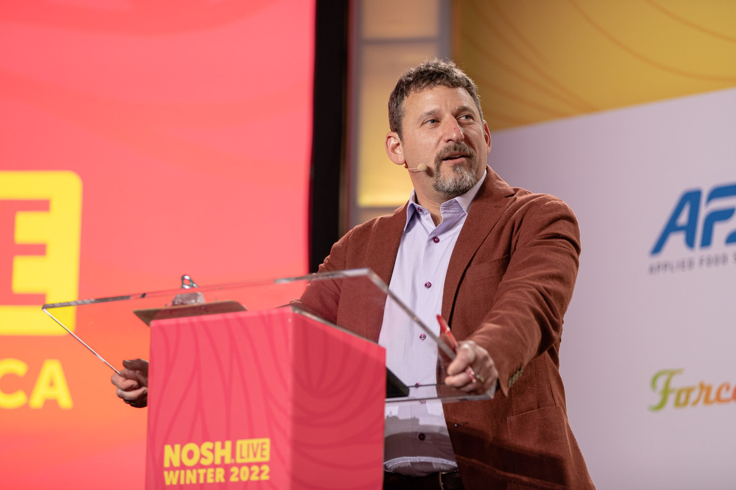 Man with a goatee in a rust blazer behind a pink Nosh Live Winter 2022 podium.