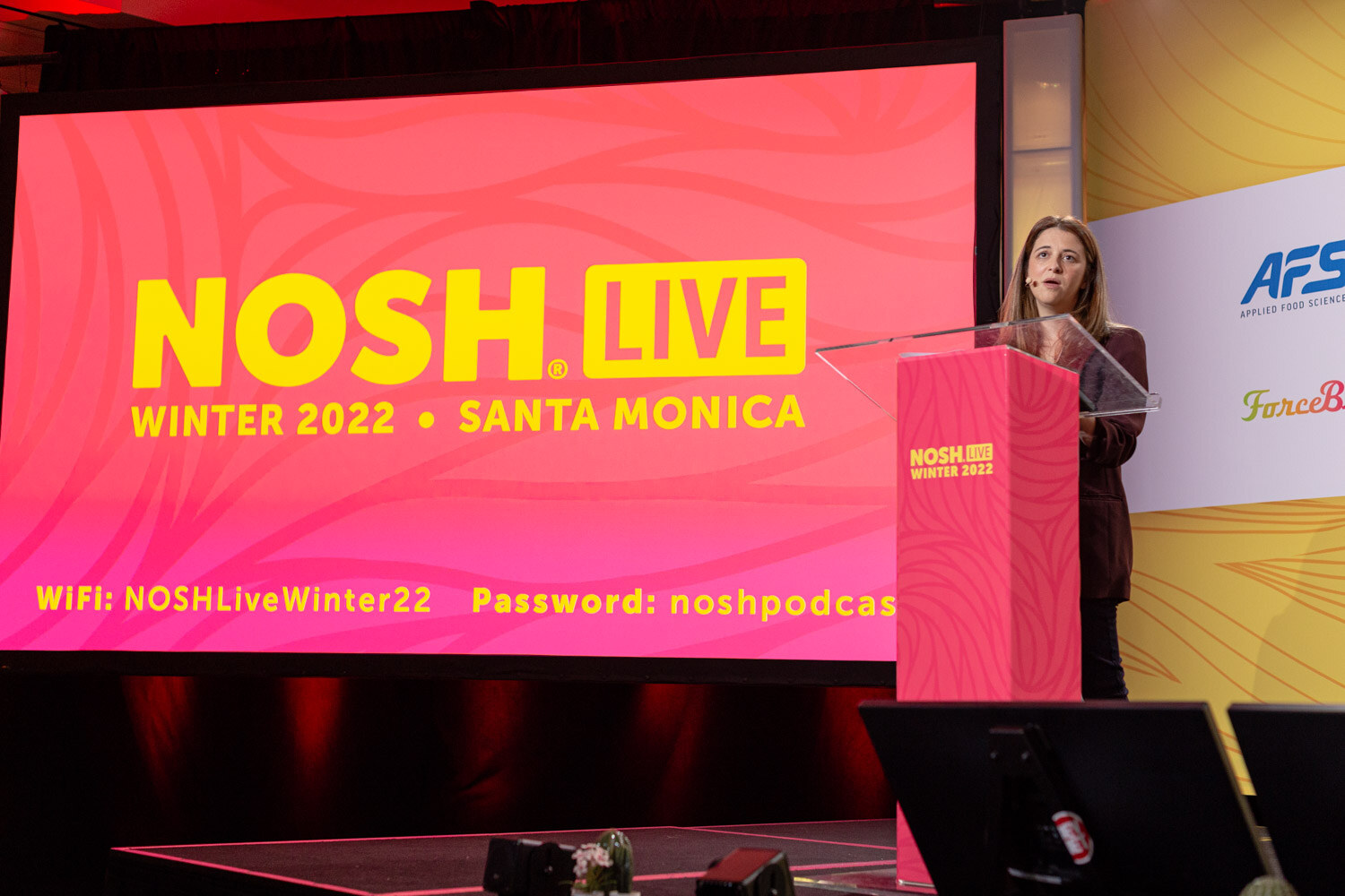 Woman at a pink lectern next to a large pink screen reading Nosh Live Winter 2022, Santa Monica.