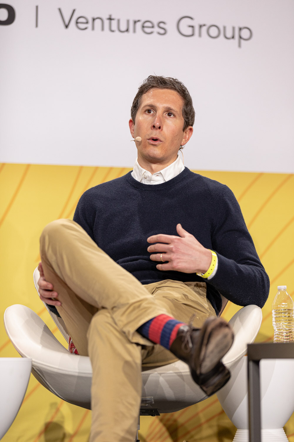 Man in a navy sweater and khaki pants seated on stage, speaking with one hand raised.