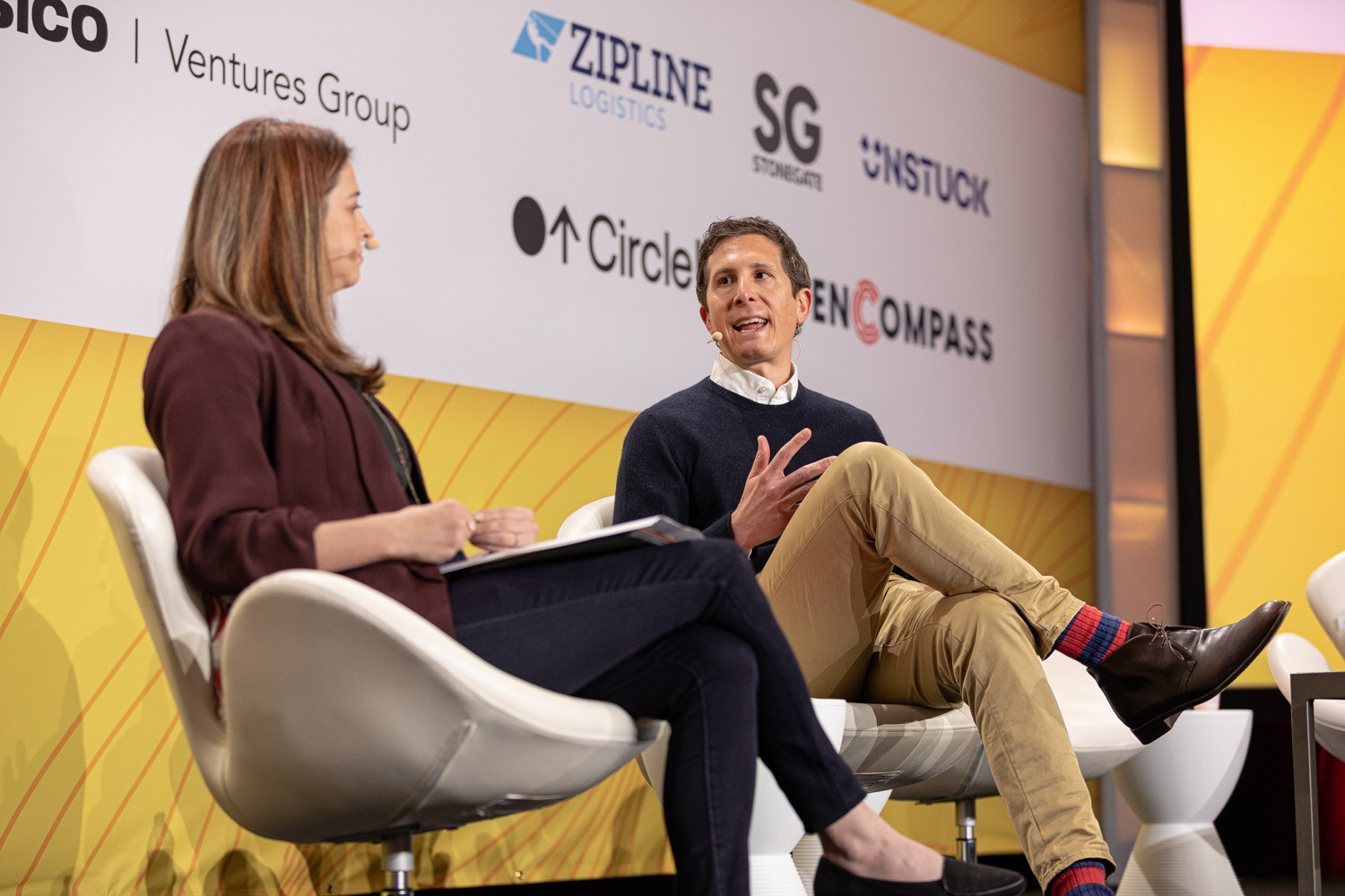 A woman holding notes and a man in a navy sweater seated on stage, the man gesturing as he speaks.