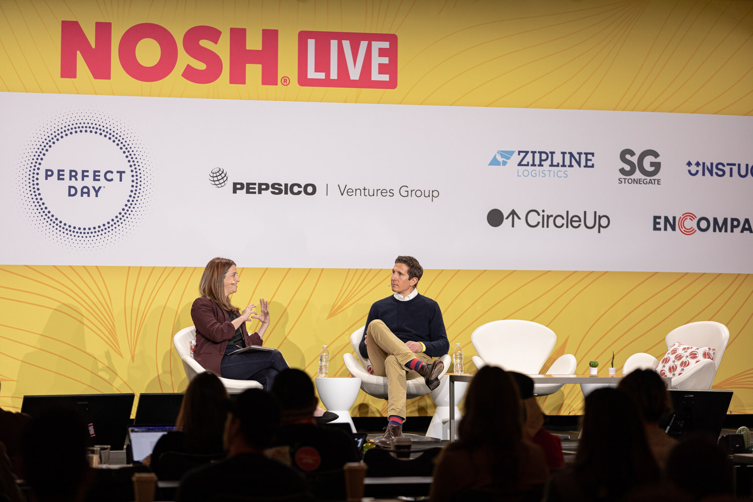 Wide shot of a stage with a Nosh Live banner, two seated speakers and audience heads in the foreground.