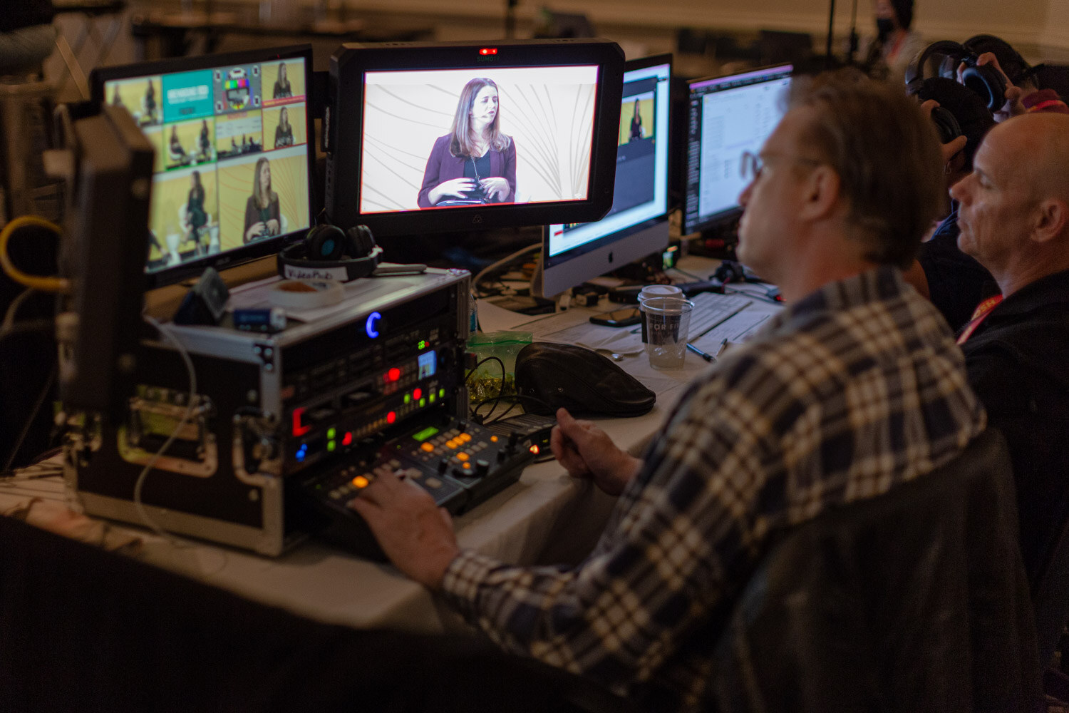 Production desk with several monitors showing a speaker on stage and a man in a plaid shirt at the controls.