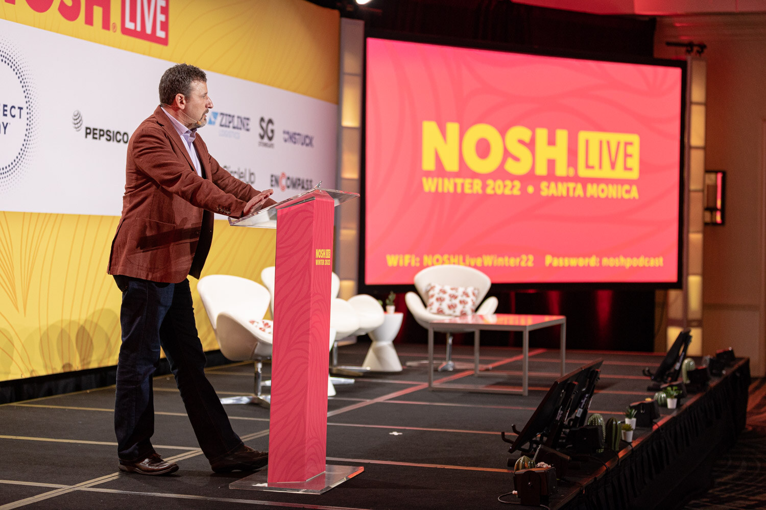 Man in a red jacket at a pink podium beside a large screen reading Nosh Live Winter 2022 Santa Monica.