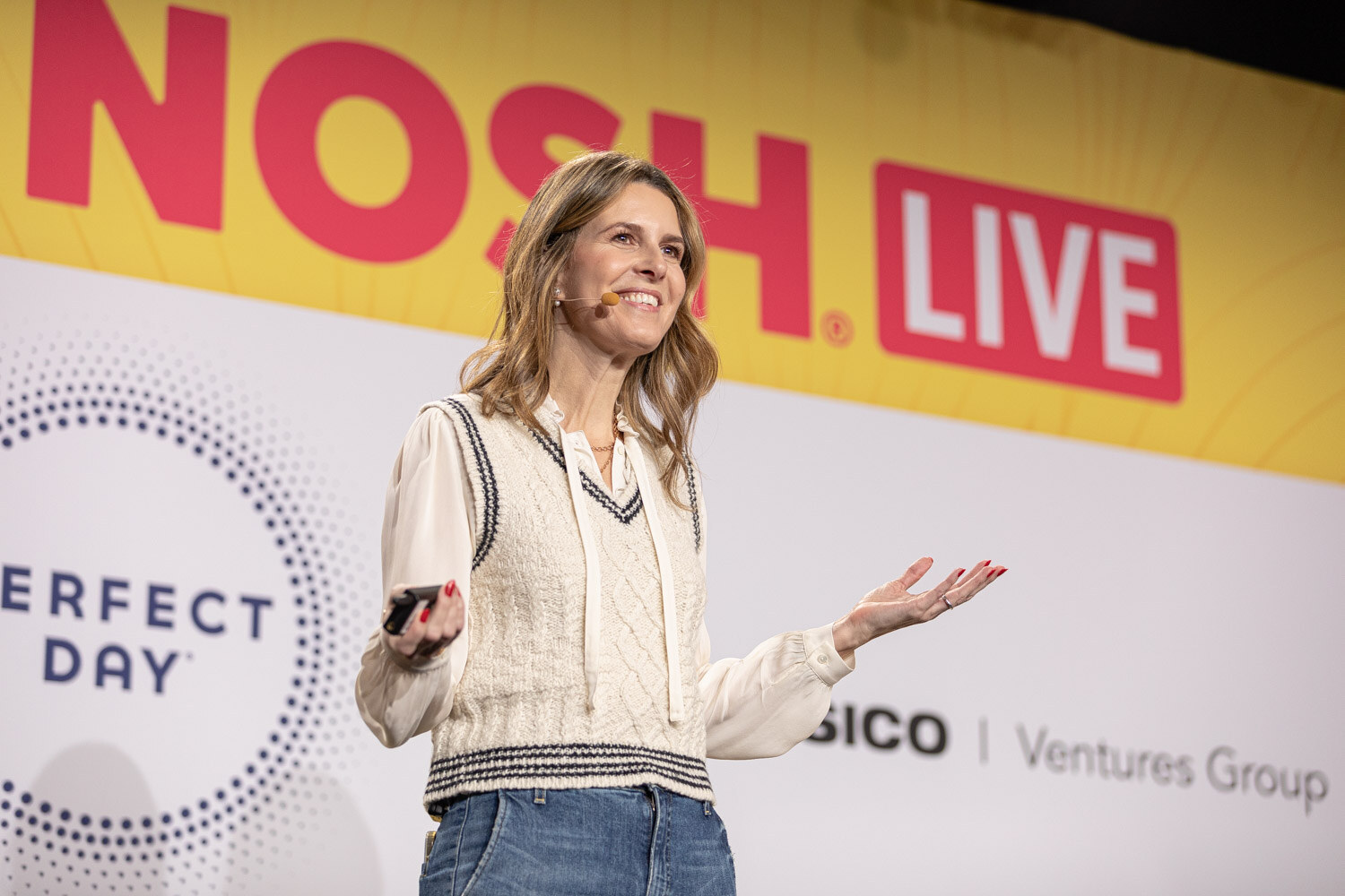 Smiling woman in a cream sweater vest holding a clicker in front of a Nosh Live banner.