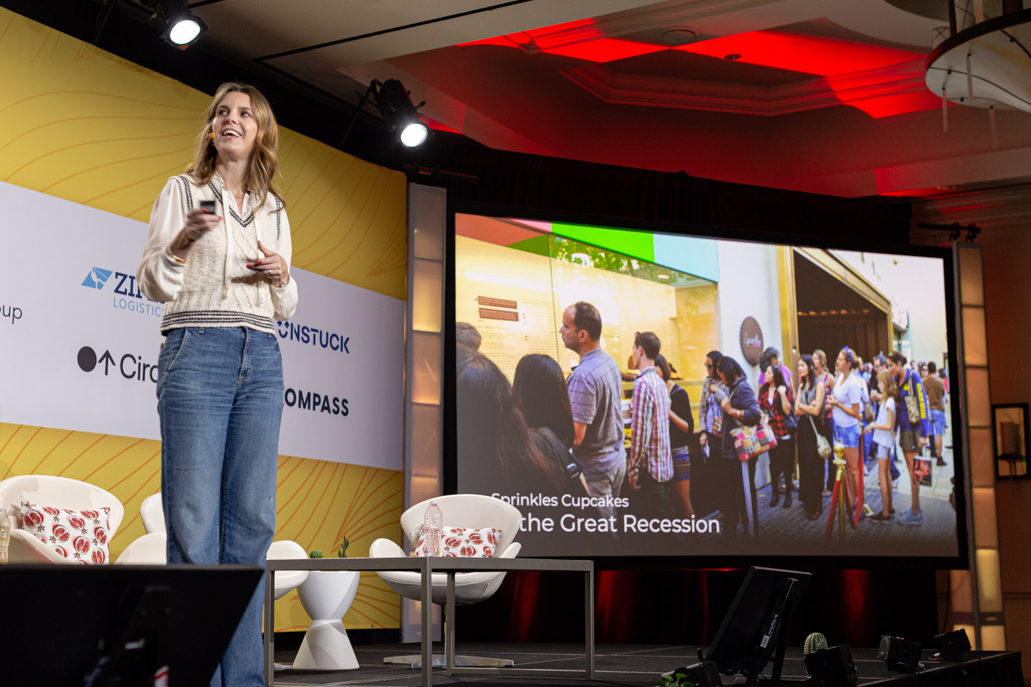 Woman presenting on stage next to a screen showing a crowd waiting in line outside a store.
