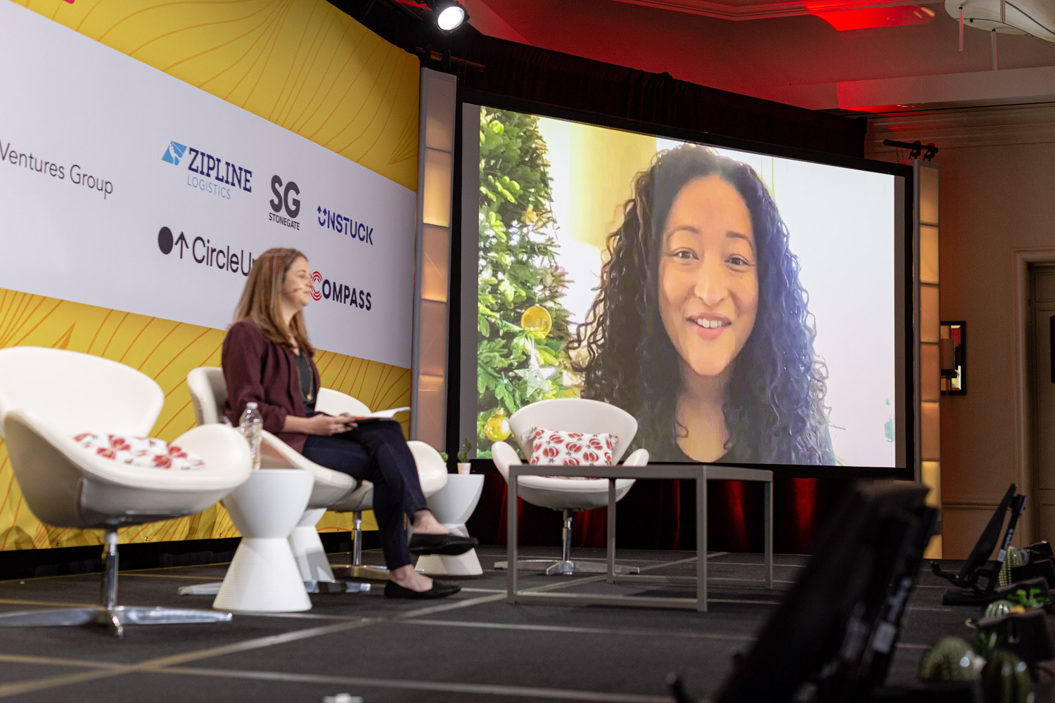 Large screen showing a smiling woman with long curly hair on a video call, a woman seated on the stage below it.