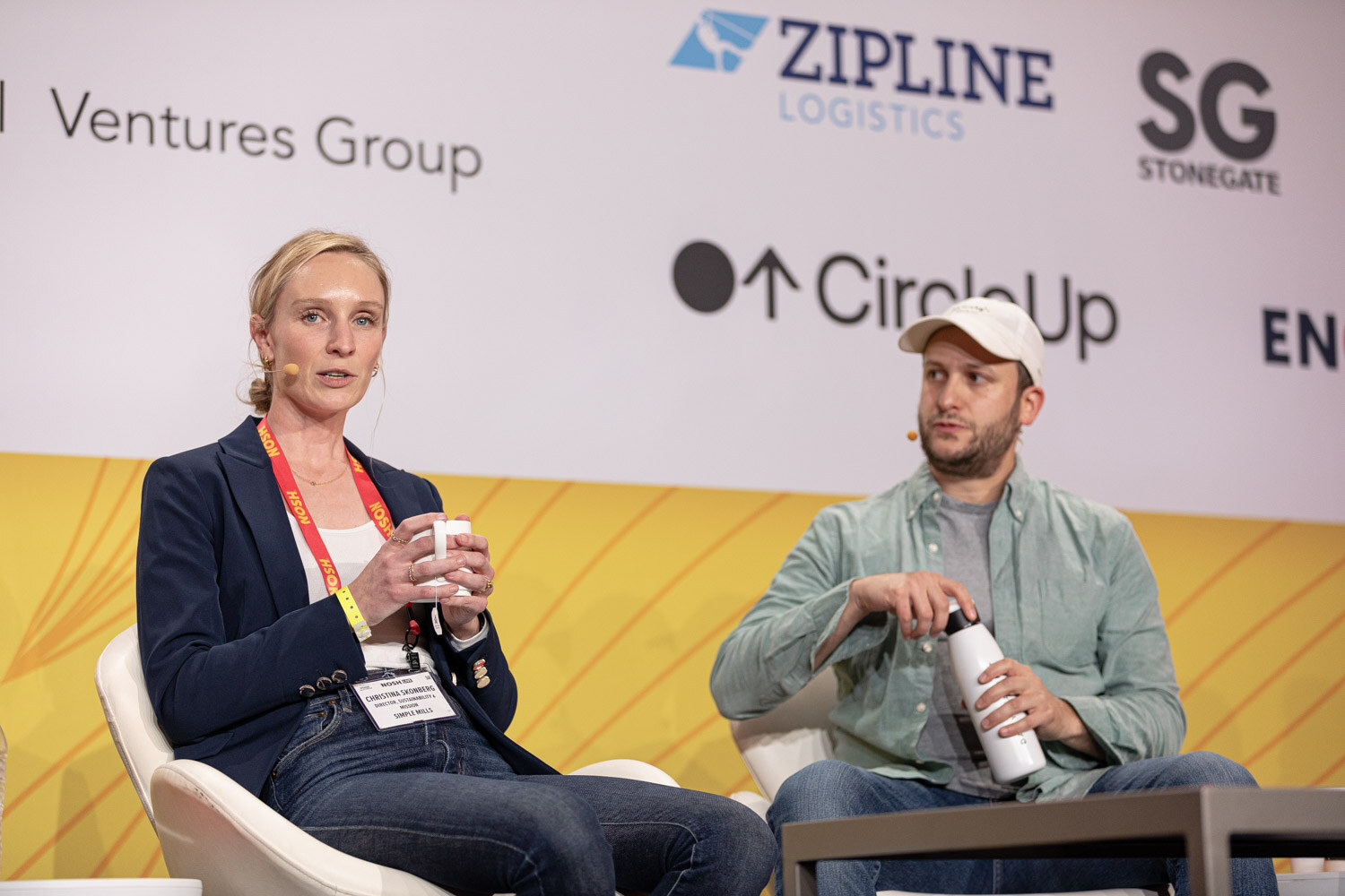 Woman in a navy blazer with a mug and man in a green shirt holding a water bottle seated on stage.