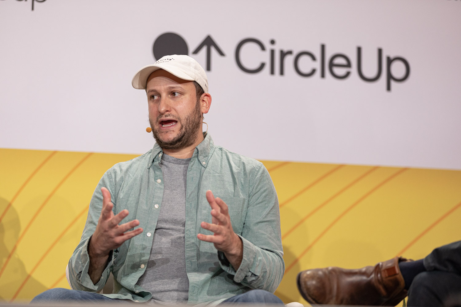 Man in a white baseball cap and green button-down shirt speaking with both hands raised.