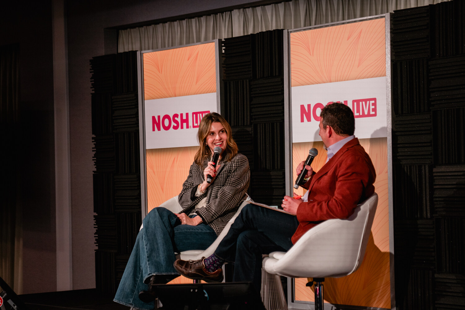 Man in a red jacket speaking into a microphone while a woman in a plaid blazer smiles at him.