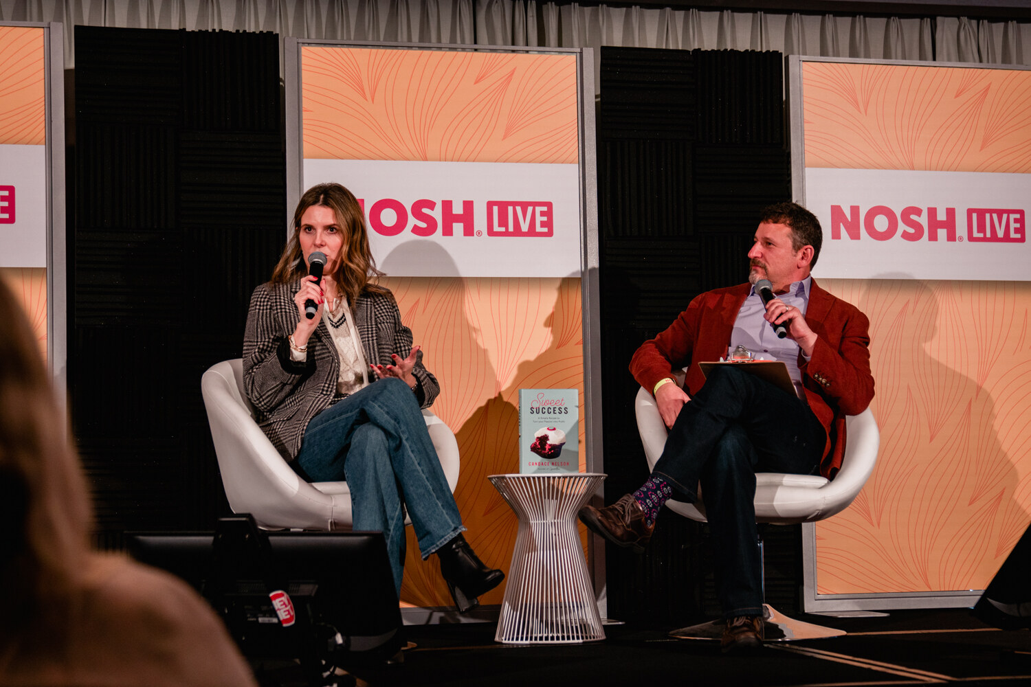 Woman in a plaid blazer and man in a red jacket seated on white chairs in front of Nosh Live panels.