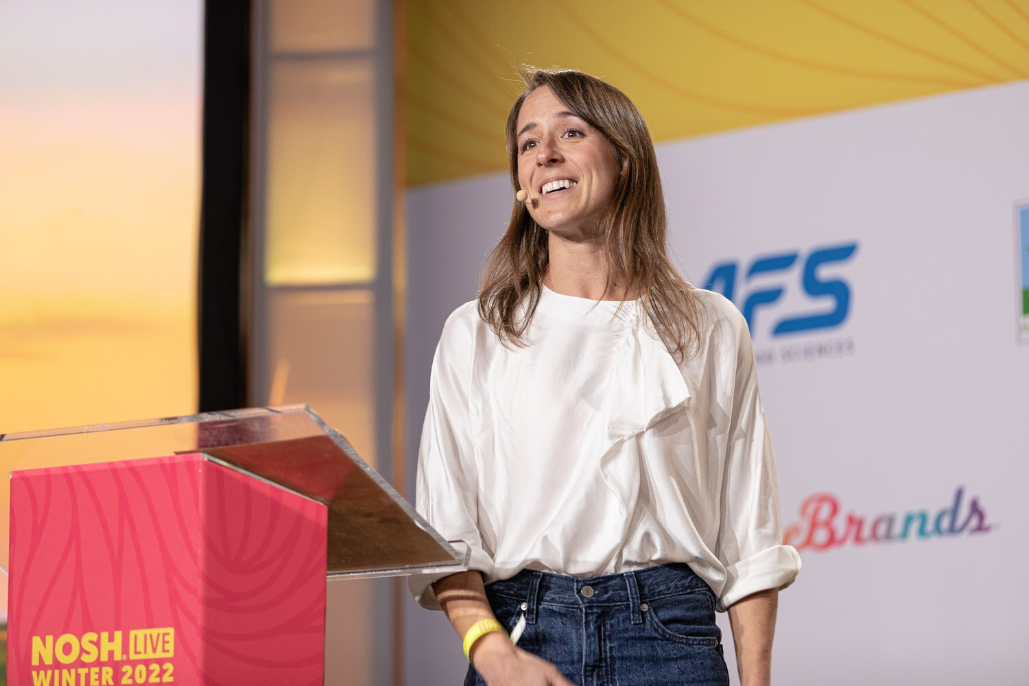 Woman in a white blouse and jeans standing beside a pink Nosh Live podium.