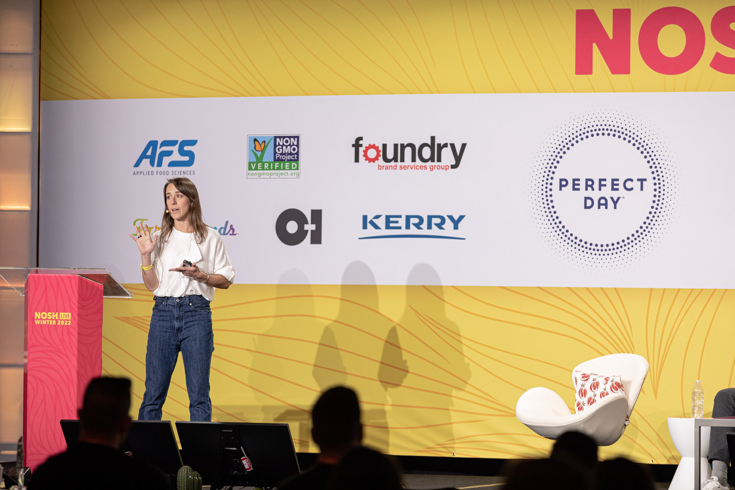 Woman in a white top standing on a stage with sponsor logos behind her and audience silhouettes in front.