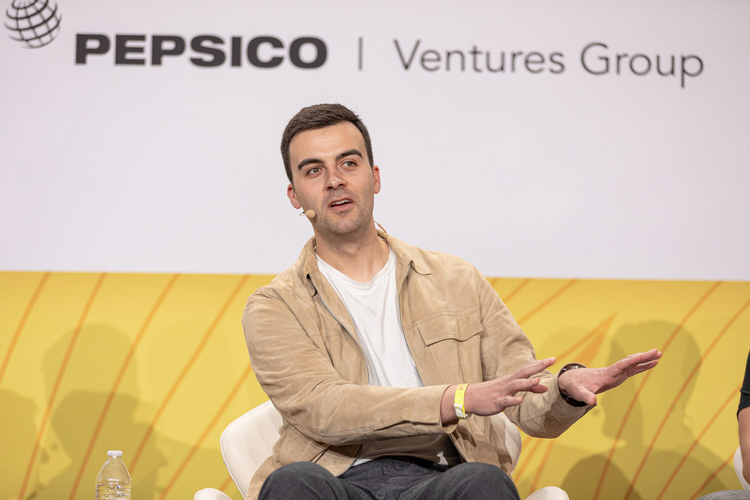 Man in a tan shirt jacket and white tee with one hand extended, in front of a Pepsico Ventures Group banner.