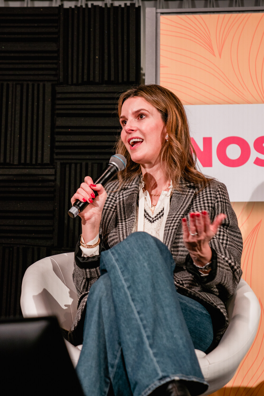 Woman in a plaid blazer and jeans gesturing while she speaks into a microphone.