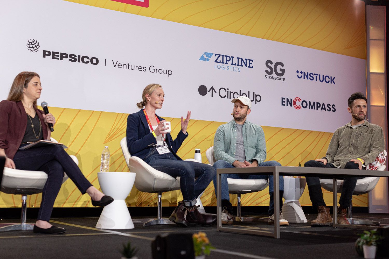 Four people seated on a stage with a sponsor wall behind them; one panelist holds a mug.
