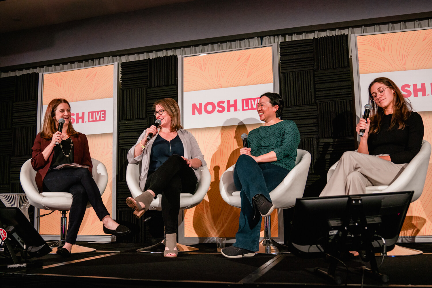 Four women seated in white chairs with microphones in front of Nosh Live panels.