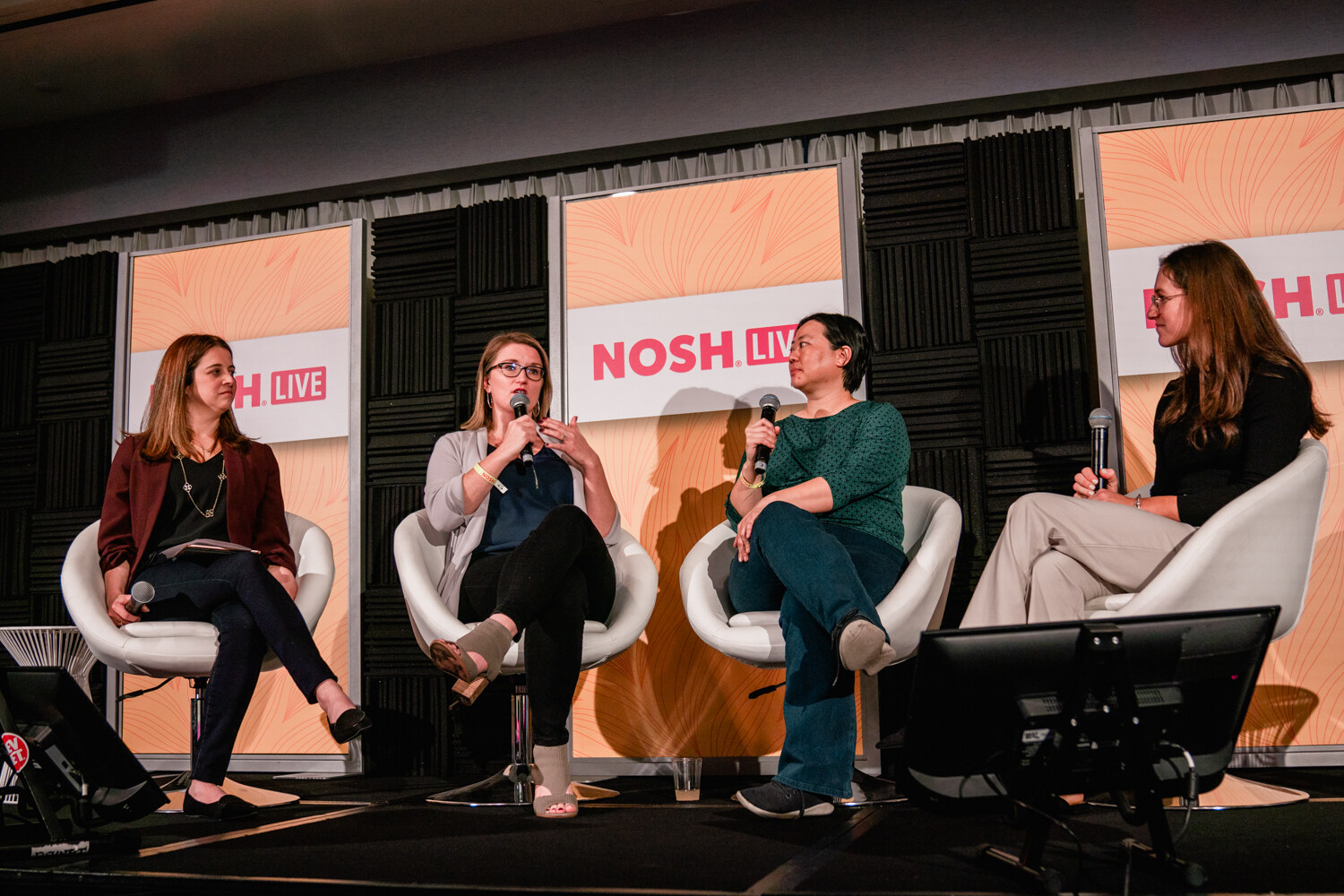 Four seated women on a stage with Nosh Live signs behind them and a monitor in the foreground.