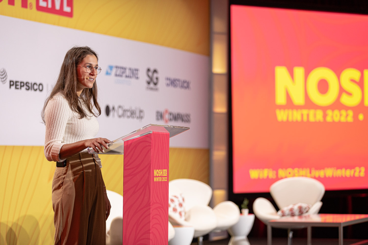 Woman in a white sweater at a pink podium with a Nosh Live Winter 2022 screen behind her.